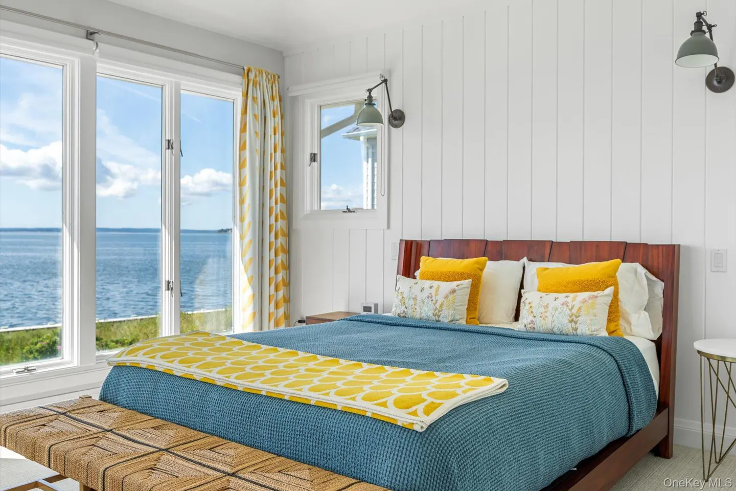 Bedroom with a blue bedspread, yellow pillows, and a view of the ocean through large windows.