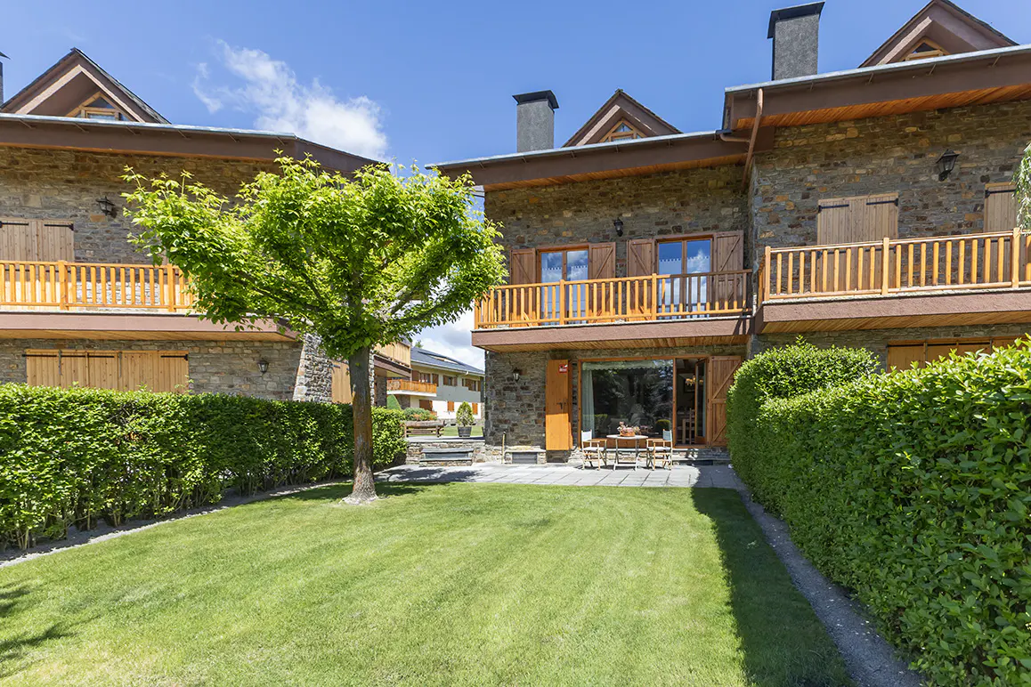 Exterior view of stone townhouses with green lawn, hedges, and a tree in the backyard on a sunny day.