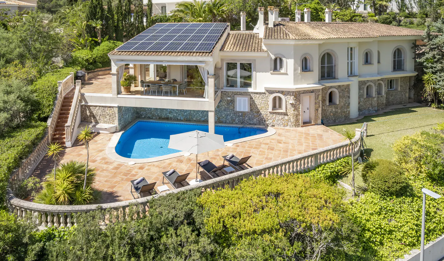 Aerial view of a two-story house with solar panels, a pool, lounge chairs, and a patio surrounded by lush greenery.