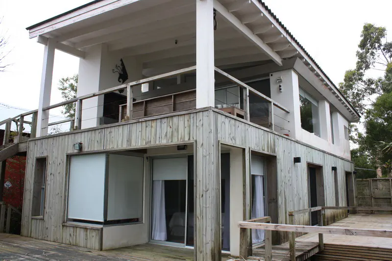 Two-story modern house with weathered wood siding, a large upper balcony, and a wooden deck in a wooded setting.