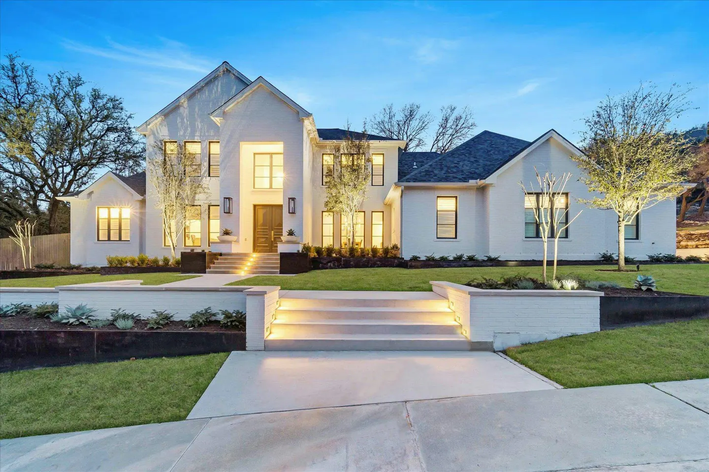 Exterior view of a two-story white house with a dark roof, illuminated windows, and a landscaped front yard.