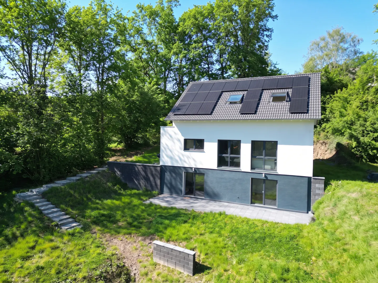 Two-story house with solar panels on a gray roof, white and gray walls, and a patio. Green grass and trees surround the house.