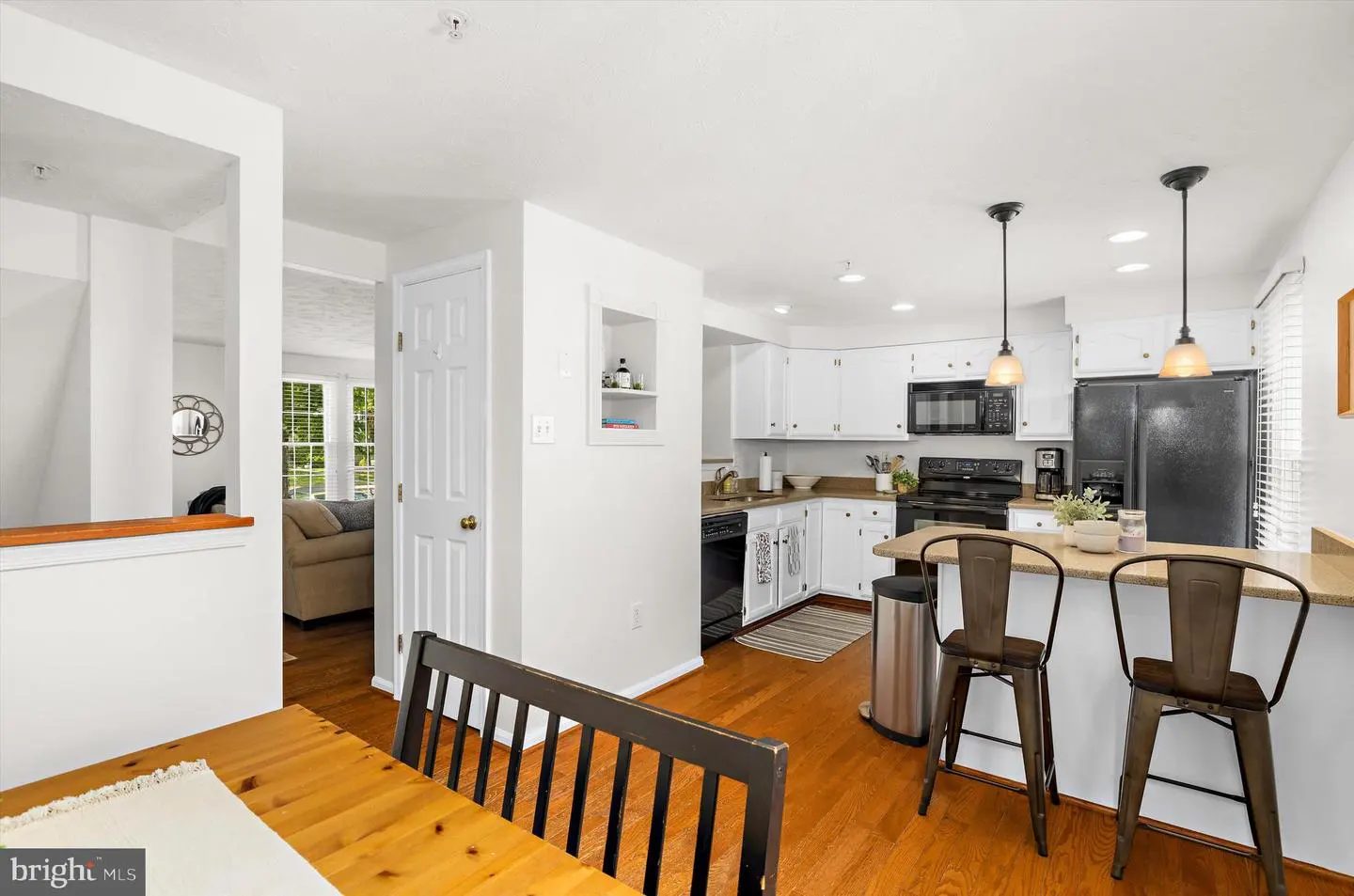 Bright kitchen with white cabinets, black appliances, and wood floors. Three bar stools line the counter. A wooden table is in the foreground.