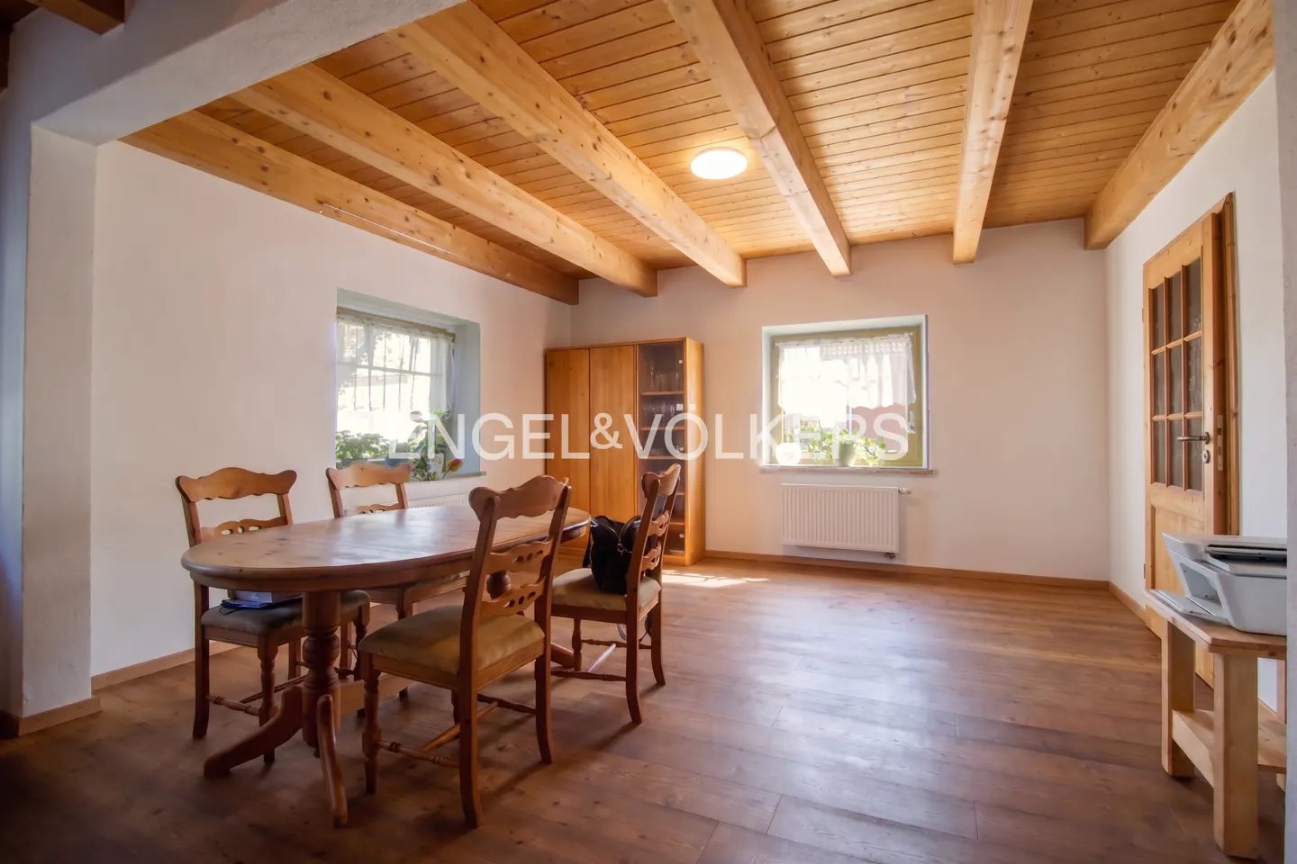 A dining room with a wooden table, chairs, and ceiling beams. Two windows let in natural light. A wooden cabinet and door add to the rustic charm.