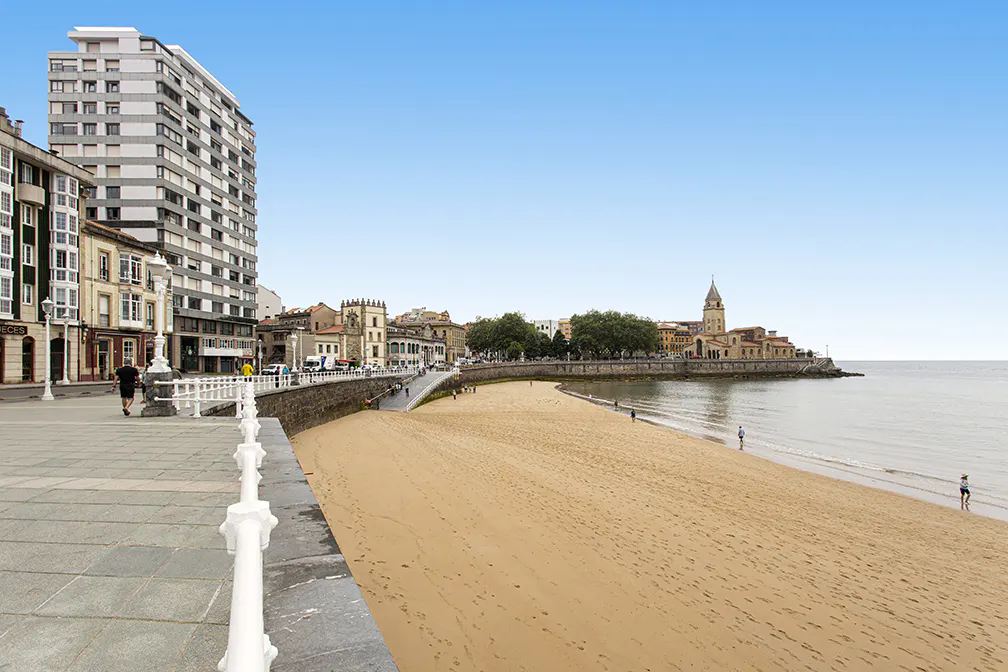 A sandy beach stretches along a coastal city promenade with tall buildings, a historic church, and a clear blue sky.
