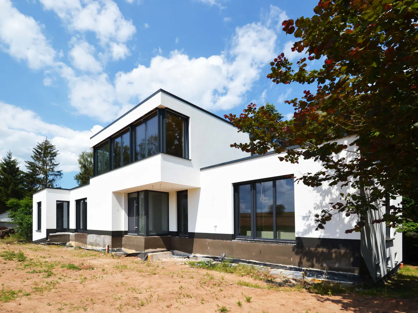Modern white house with black trim and large windows under a blue sky with scattered clouds. A tree with red leaves is on the right.