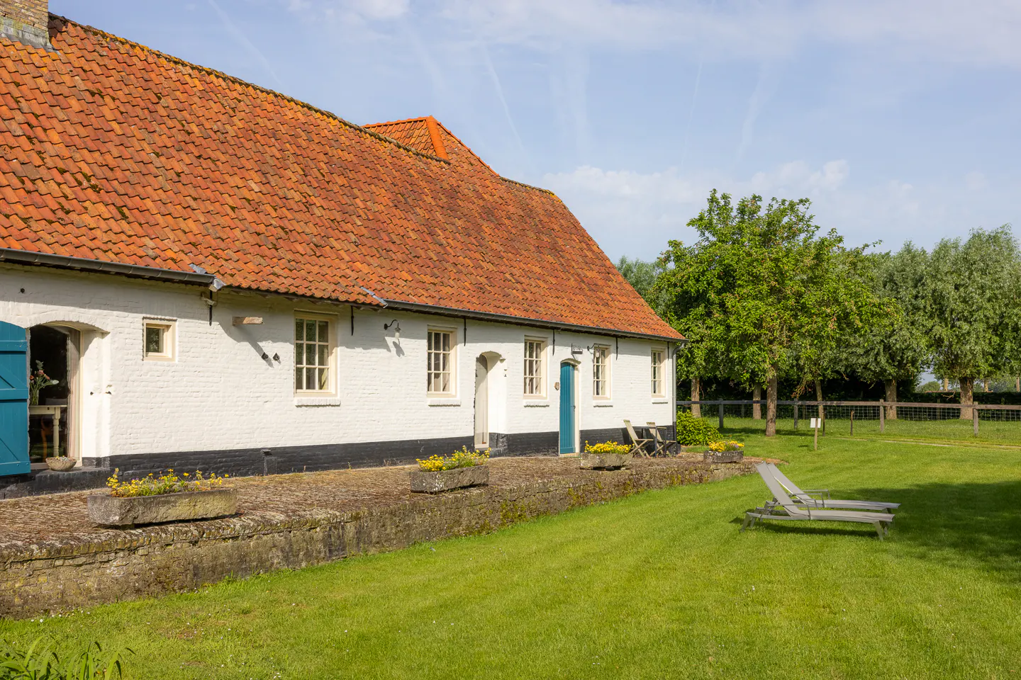 White farmhouse with a red tiled roof, stone patio, flower boxes, and lounge chairs on a green lawn under a blue sky.