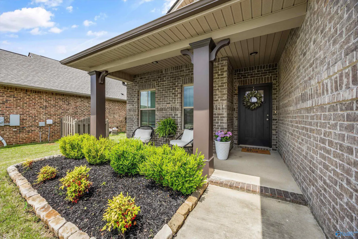 Brick home exterior with covered porch, dark brown columns, and black front door with wreath. Green bushes line the front of the house.