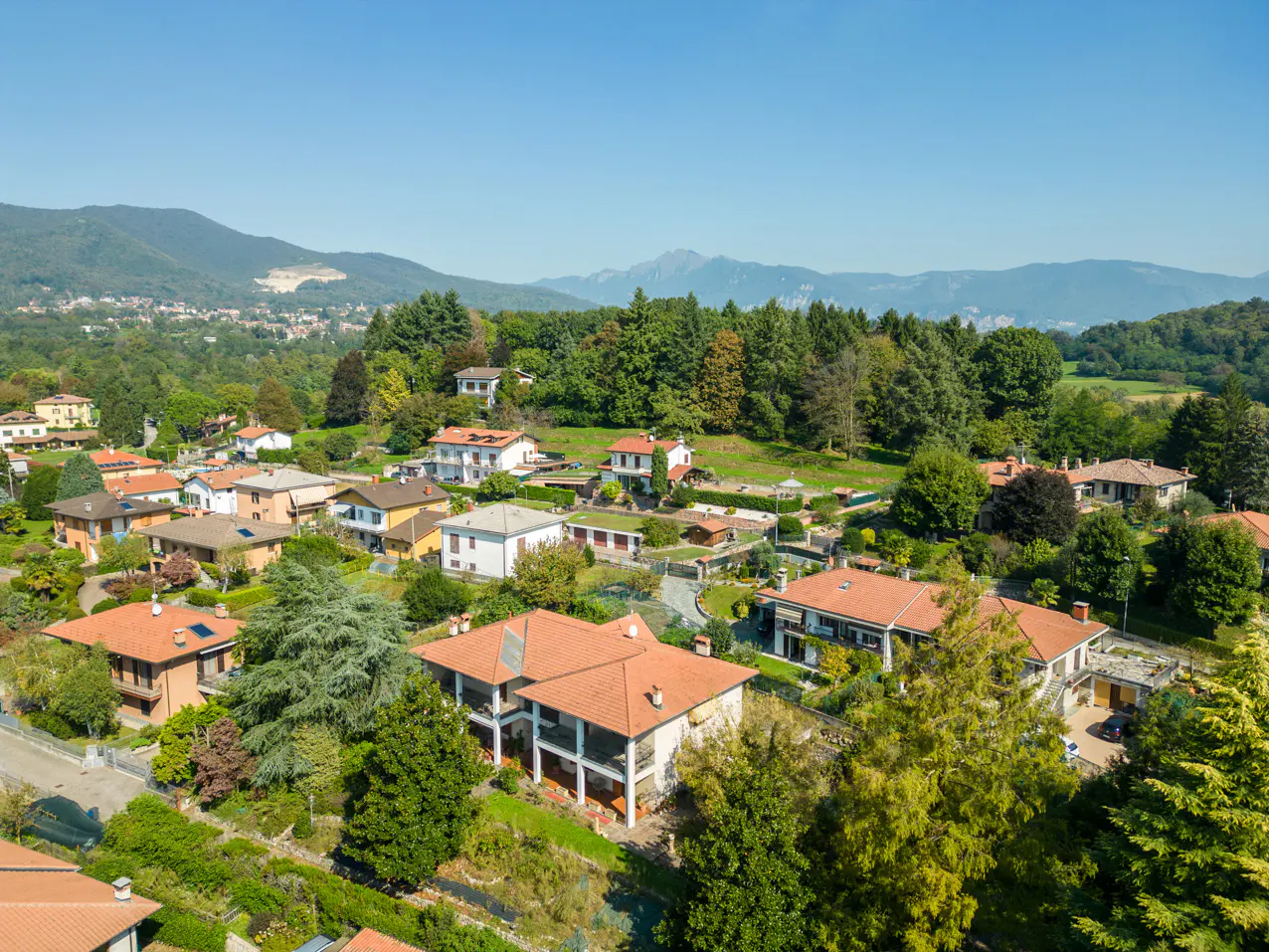 Aerial view of a residential area with houses with red tile roofs, green trees, and mountains in the background under a blue sky.