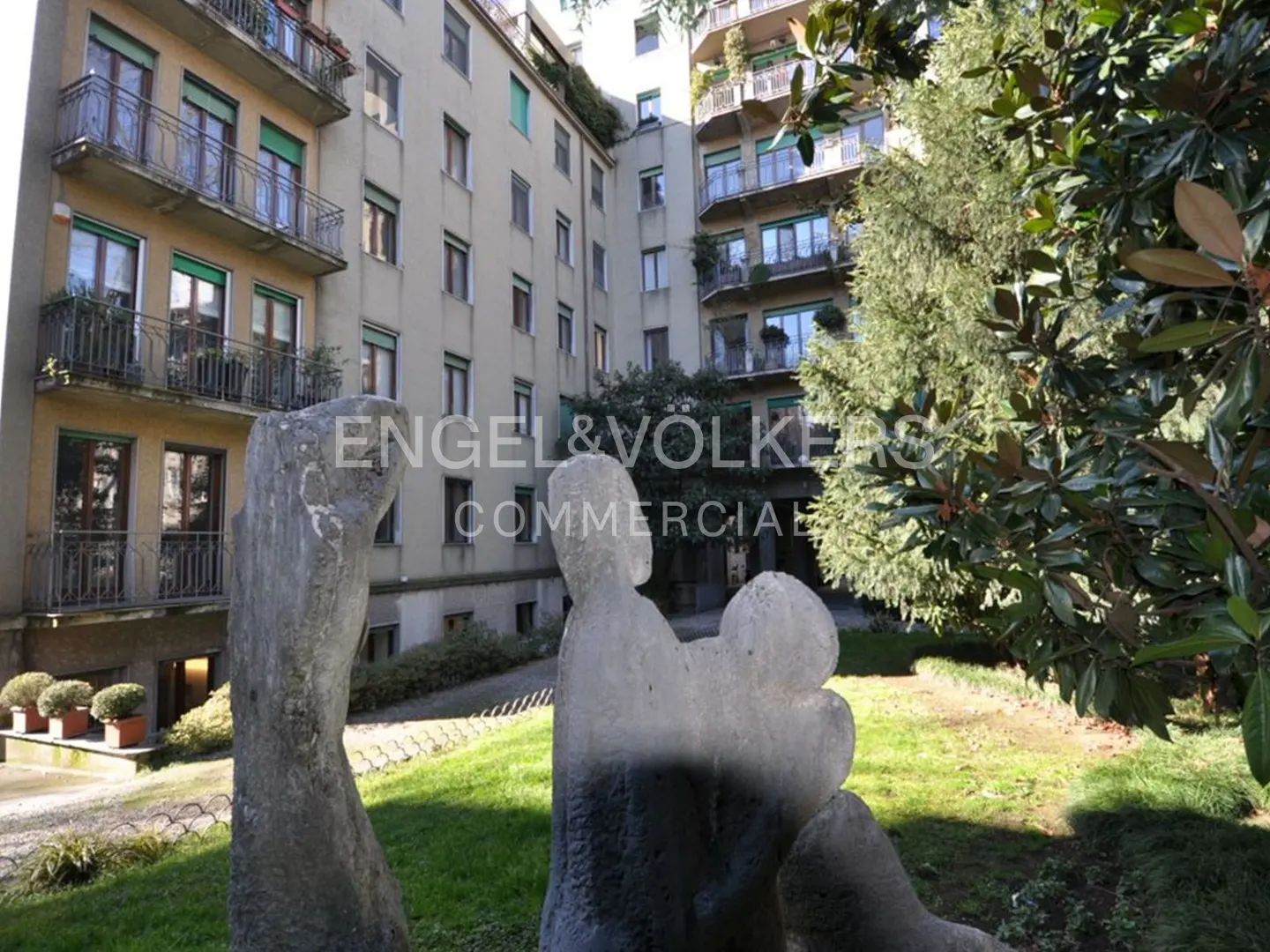 Apartment building with balconies and green window shades, seen from a garden with stone sculptures and the Engel & Volkers logo.