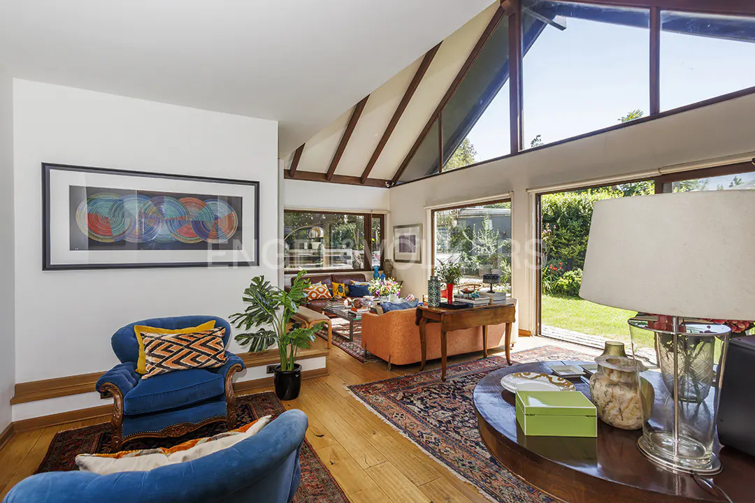 Bright living room with hardwood floors, blue armchairs, and a large window overlooking a green garden. Artwork hangs on the white walls.