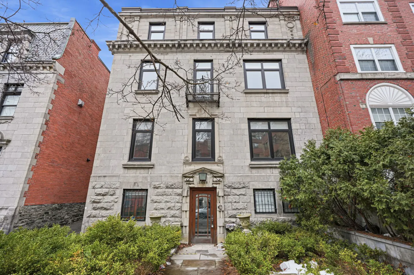 Exterior view of a 5-story gray stone building with black-framed windows and a brown front door. Green bushes line the front.