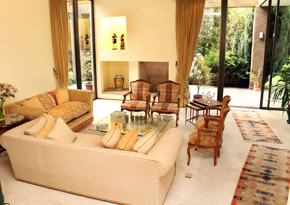 A bright living room with sofas, chairs, and a glass table. Large windows overlook a lush green garden.