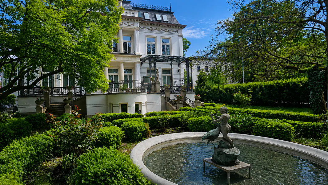 Elegant white mansion with manicured green gardens and a fountain with a cherub statue in the foreground.