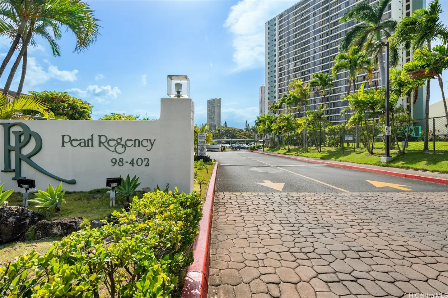 Entrance sign for Pearl Regency apartments with a stone driveway, palm trees, and a tall residential building in the back.