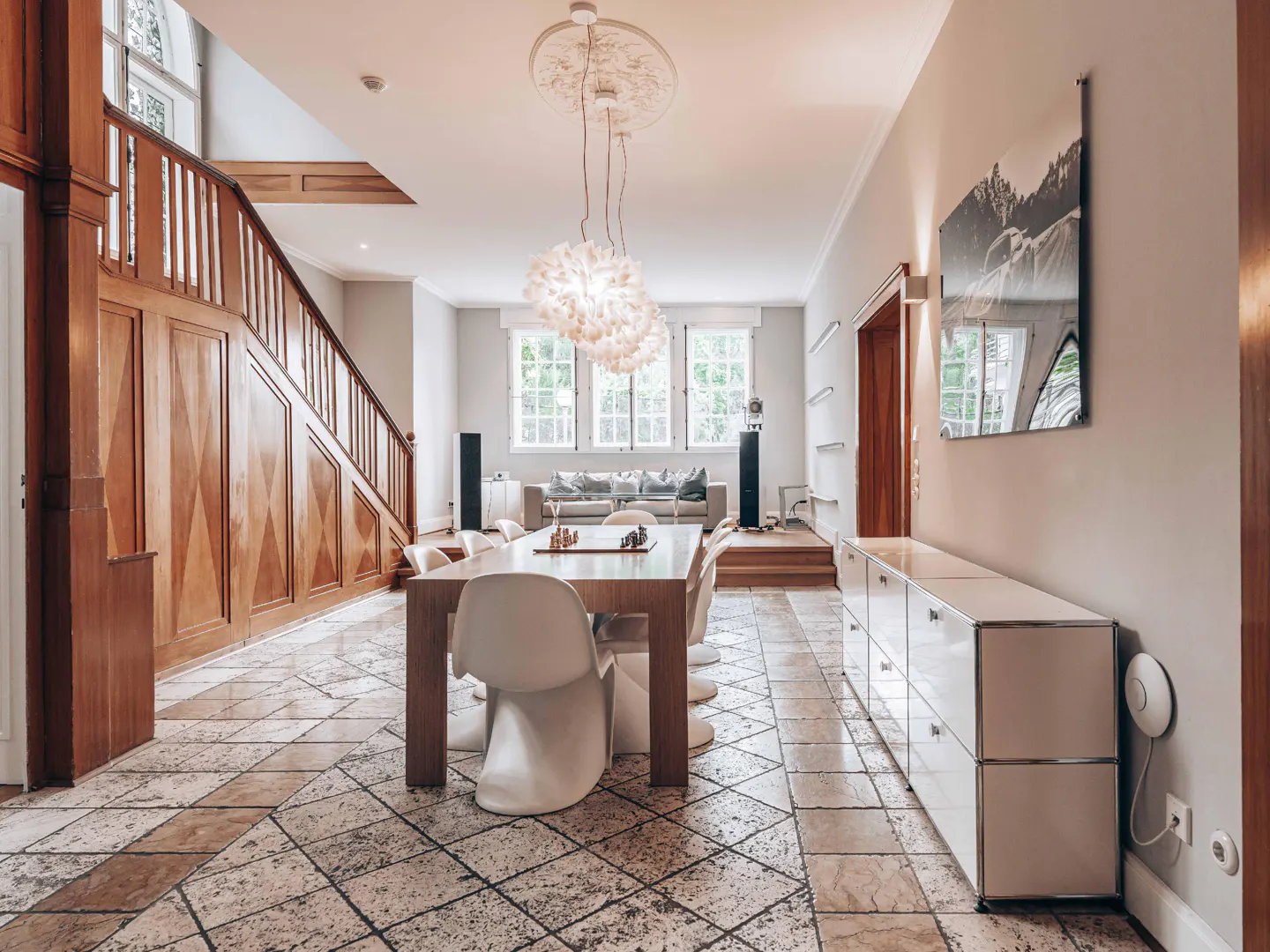 A spacious room with a dining table, white chairs, and a wooden staircase. A white cabinet is on the right, and a sofa is visible in the background.