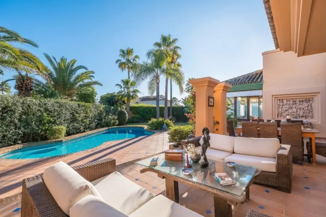 Outdoor patio with wicker furniture, a glass table, and a pool surrounded by palm trees on a sunny day.