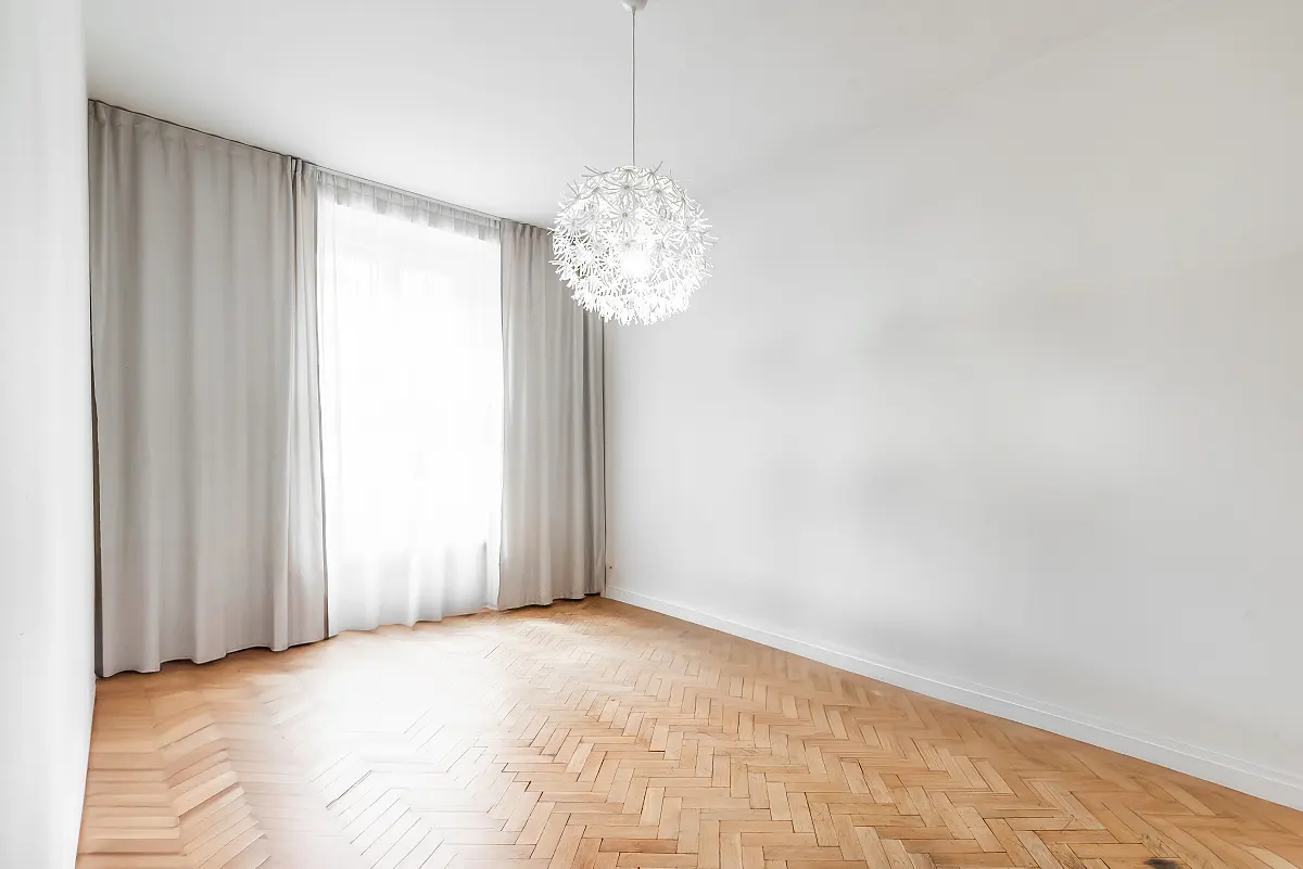 Bright, empty room with herringbone wood floors, white walls, and a modern, dandelion-shaped light fixture. Gray curtains frame a window.