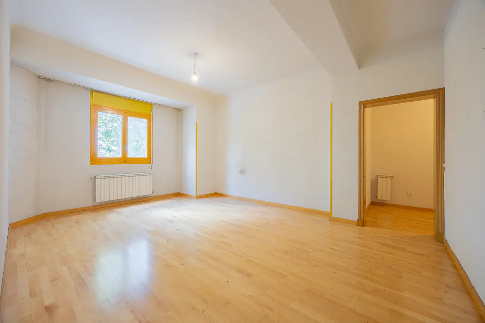 Empty room with light wood floors, white walls, and a yellow-framed window. A doorway leads to another room with a radiator.