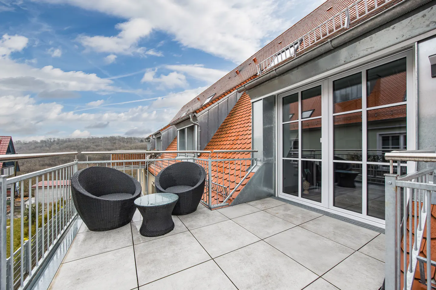 Balcony with two black wicker chairs, a small table, and a metal railing. Tiled floor, large windows, and a view of trees and sky.