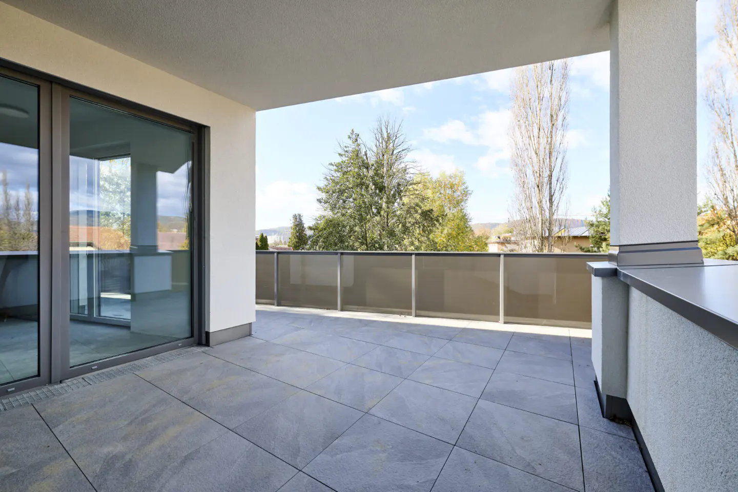 Spacious modern balcony with grey tiled flooring, glass railings, and a view of trees under a bright blue sky.