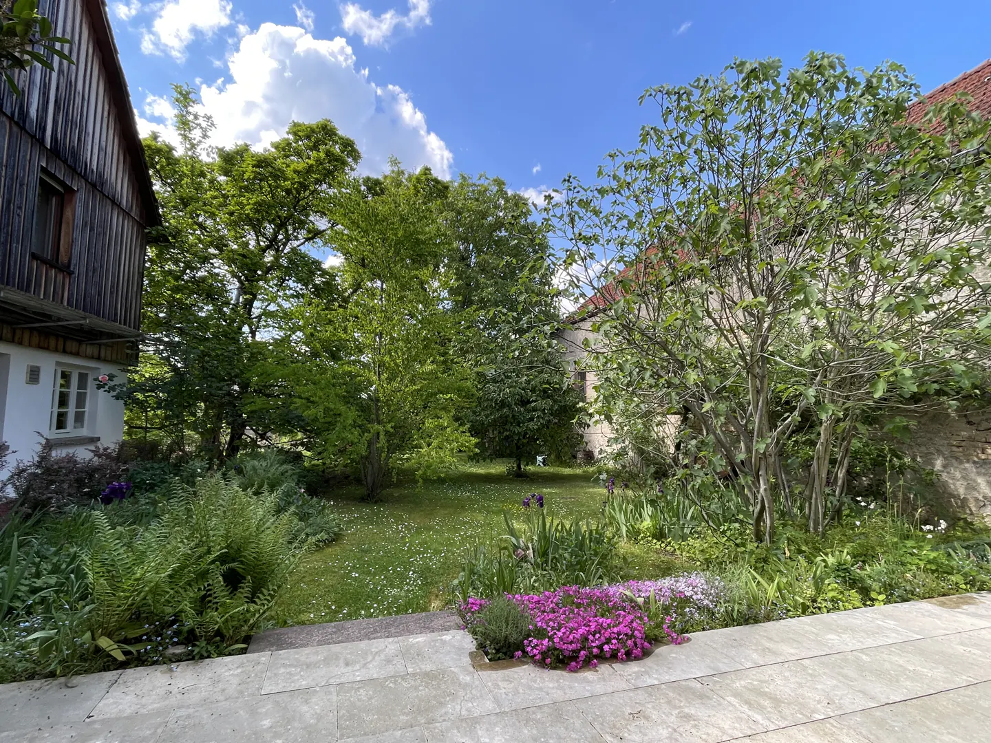 A lush garden with green grass, trees, and colorful flowers, next to a wooden house under a blue sky with clouds.