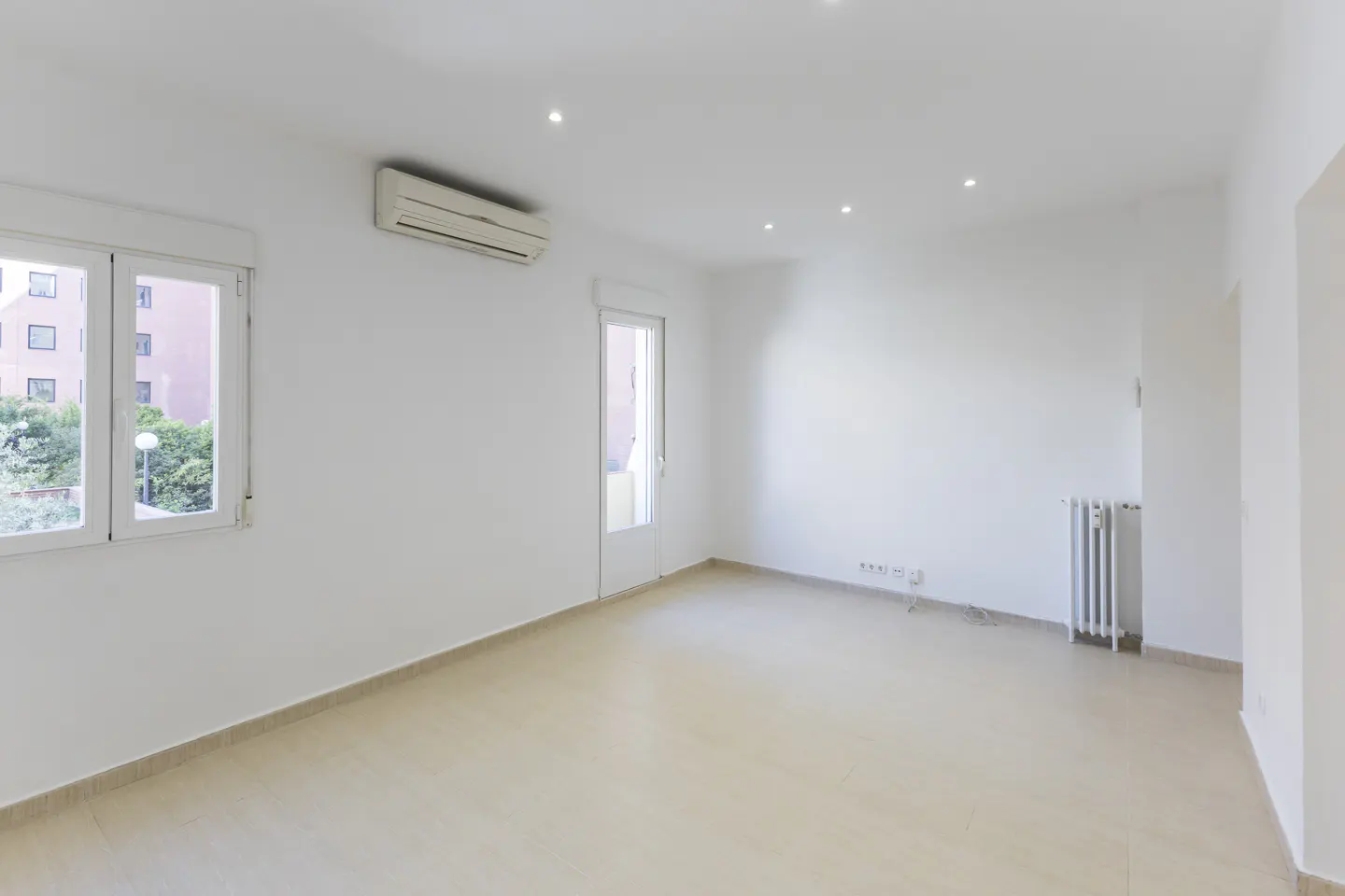 Bright, empty room with white walls, beige tile floor, window, door, AC unit, and radiator.