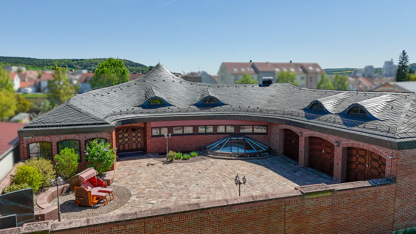 Aerial view of a red brick house with a gray shingled roof, arched windows, and a brick-paved courtyard with a red and white striped beach chair.