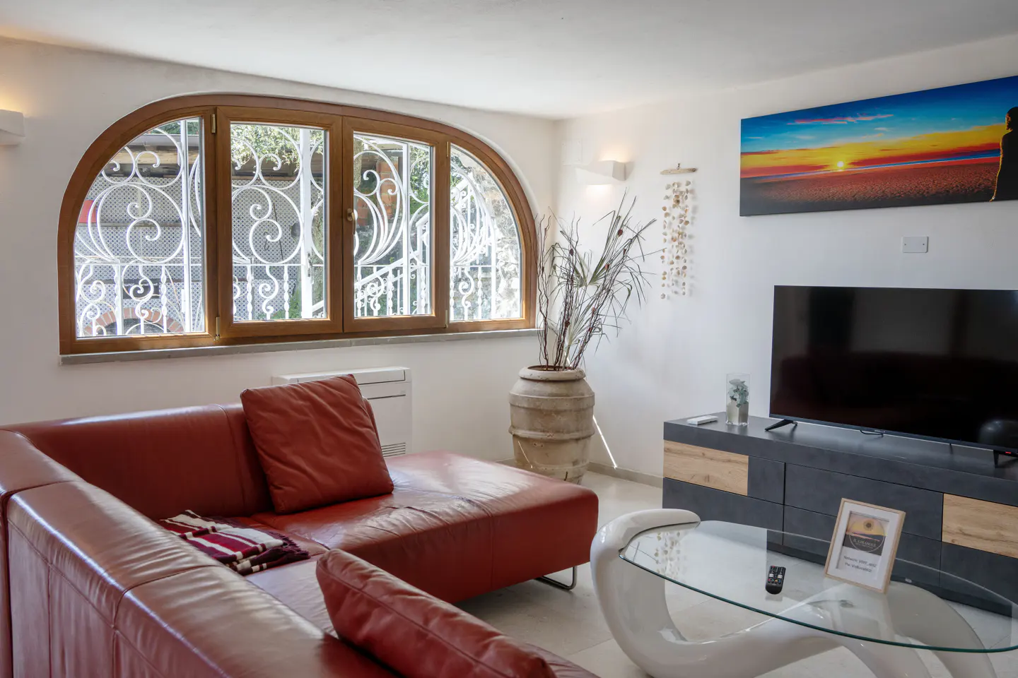 Living room with a red leather sofa, arched window with white ironwork, TV, and glass coffee table.