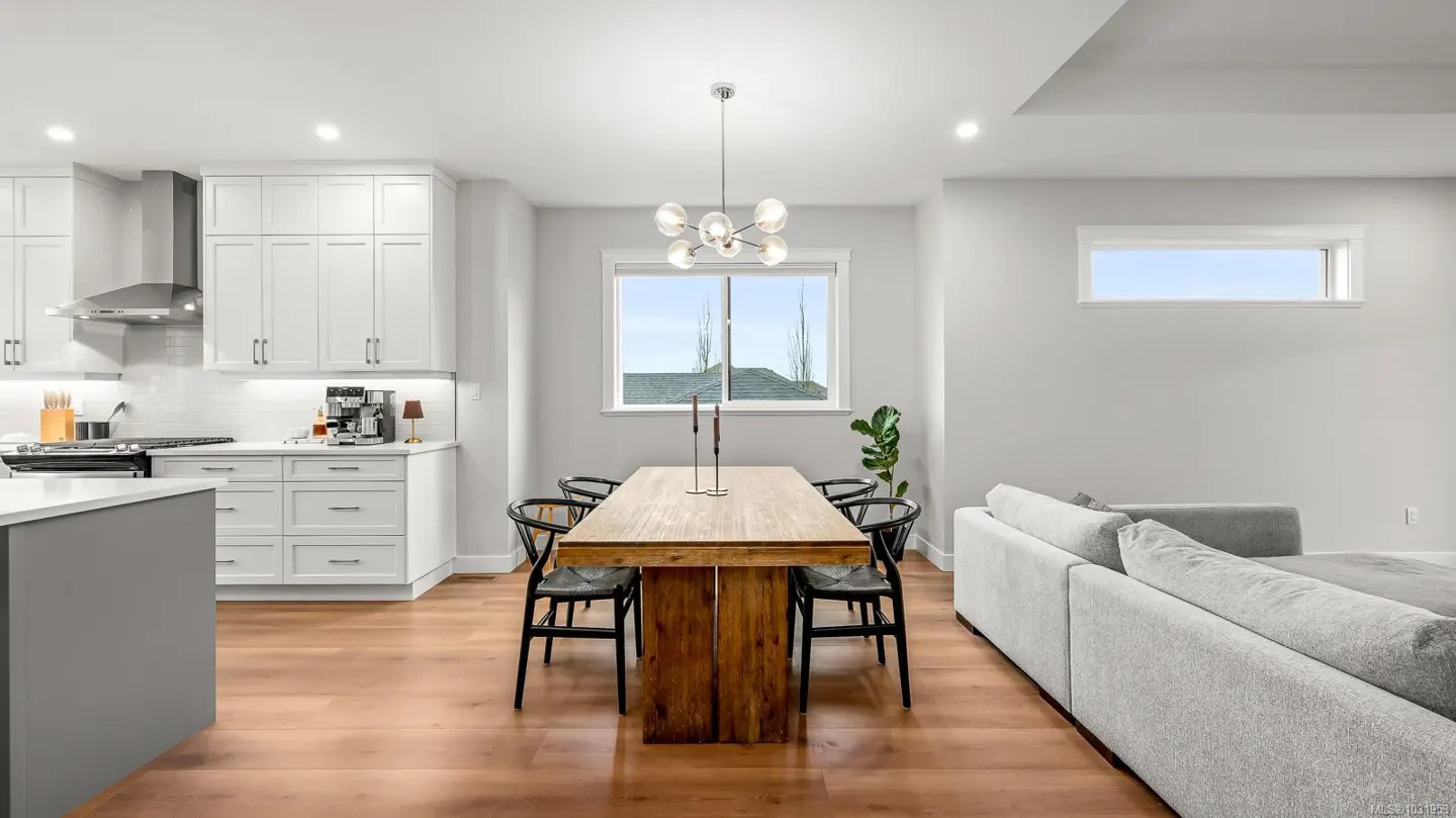 A bright, modern open-concept kitchen and dining area with wood floors. White cabinets, a wood table with black chairs, and a gray sofa are visible.