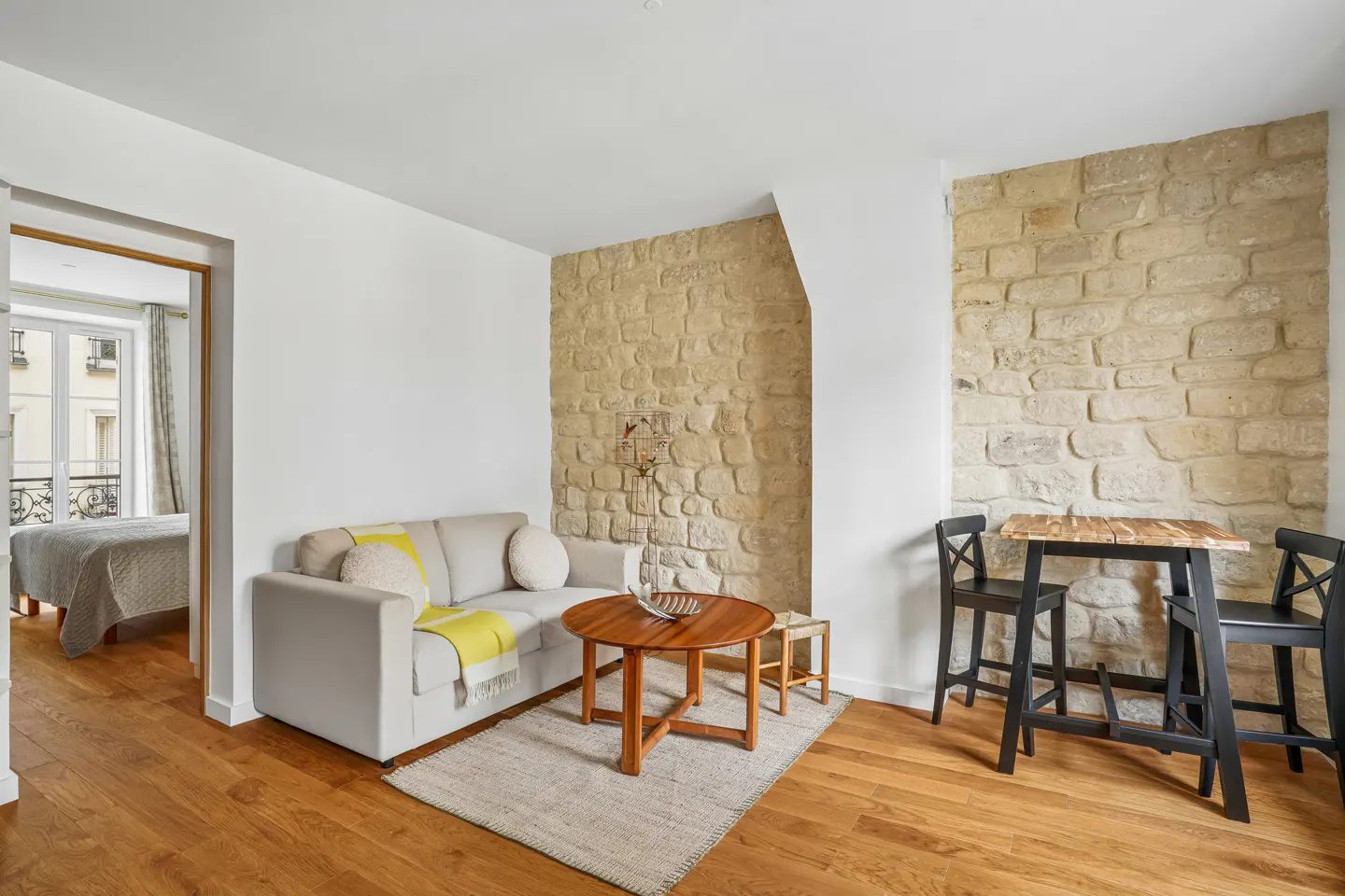 Living room with stone walls, wood floors, and a gray sofa with a yellow throw. A round wood table sits on a gray rug. A bedroom is visible through a doorway.