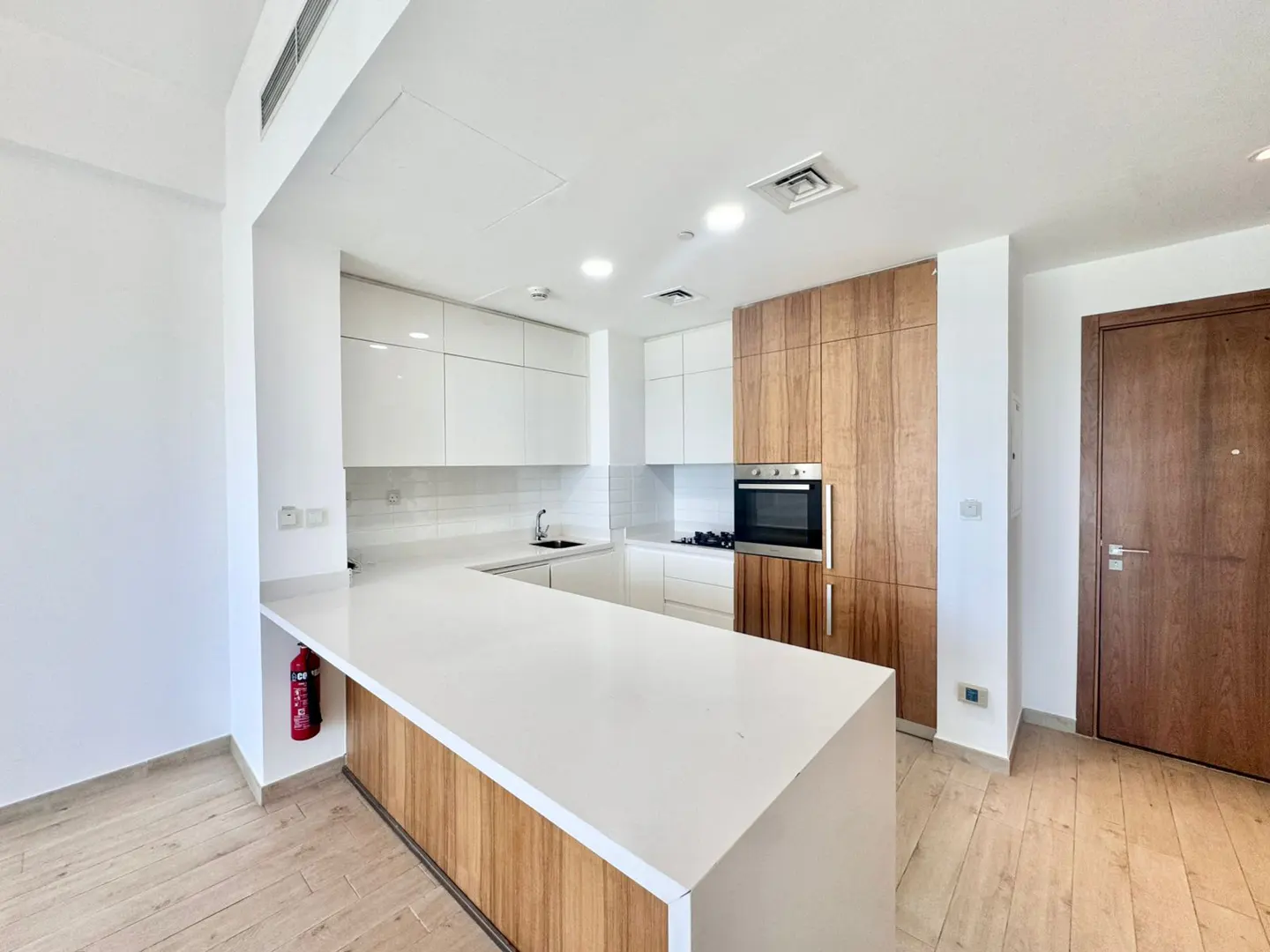 A modern kitchen with white cabinets, wood accents, and a white countertop island. A stainless steel oven is built into the wood cabinetry. A red fire extinguisher is visible.