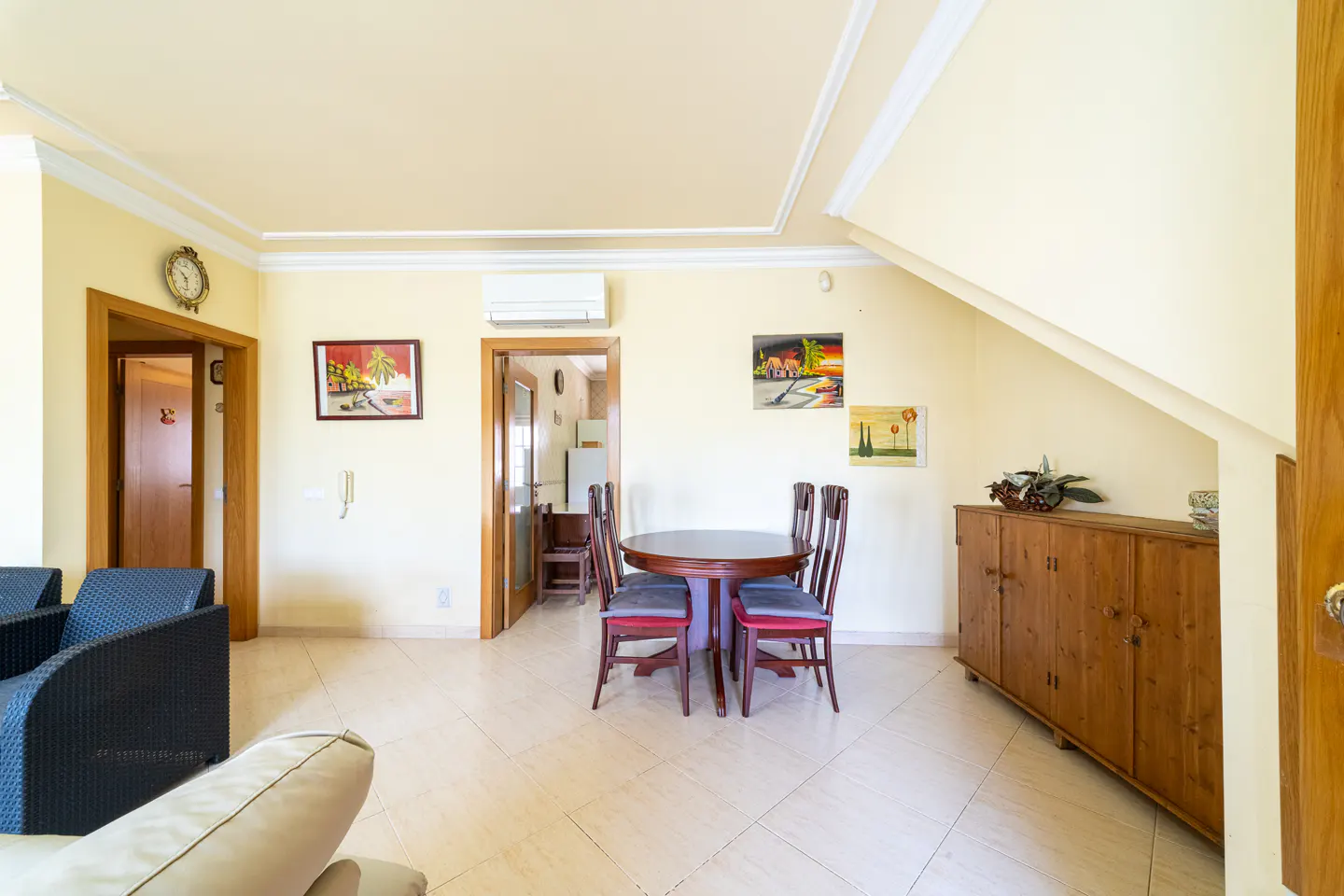 A bright living room with a round wooden table and four chairs, a wooden cabinet, and tiled floors. Paintings adorn the yellow walls.