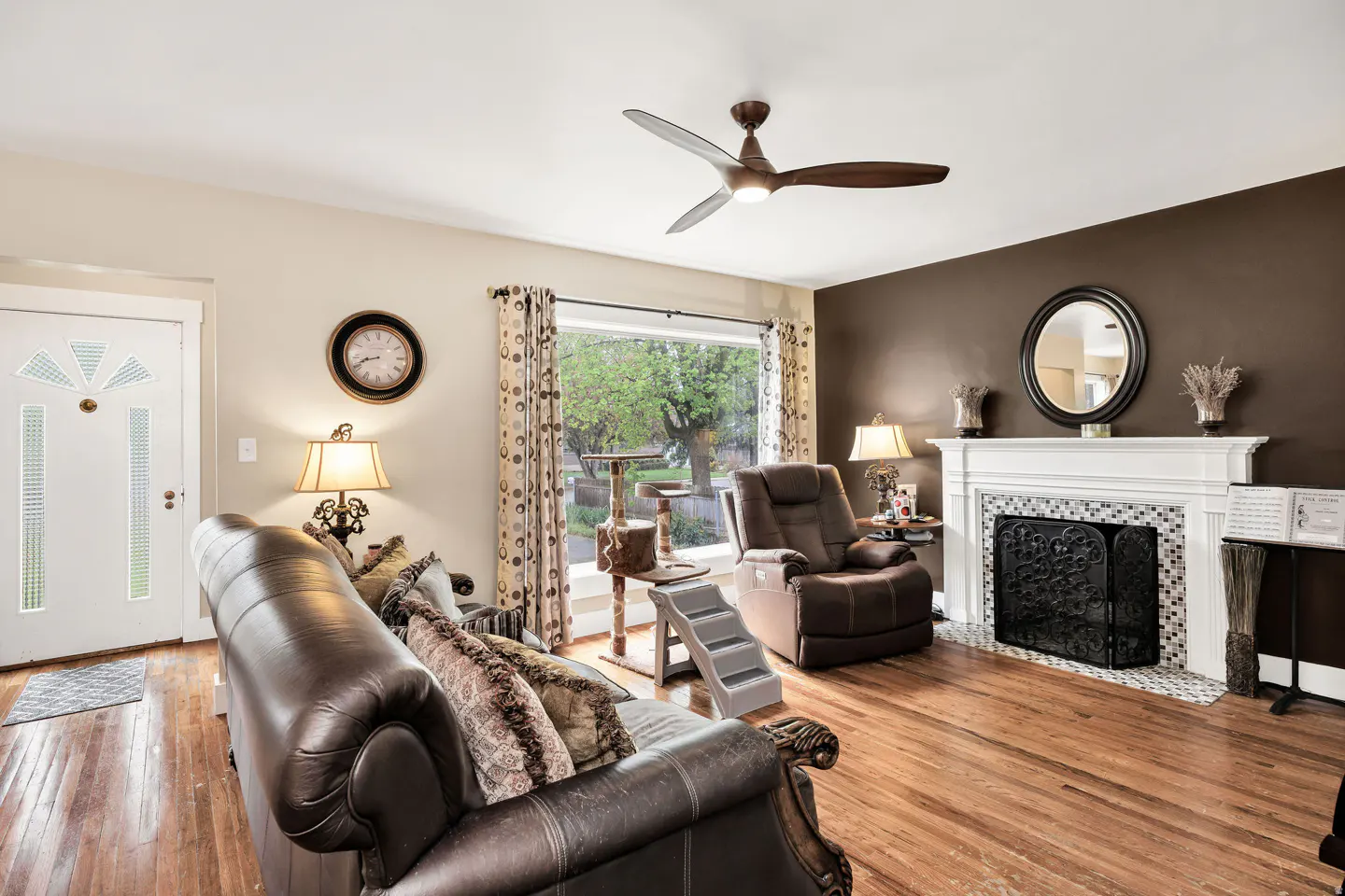 Living room with brown leather sofa, recliner, fireplace, and a cat tree near a window. Hardwood floors and a ceiling fan are visible.