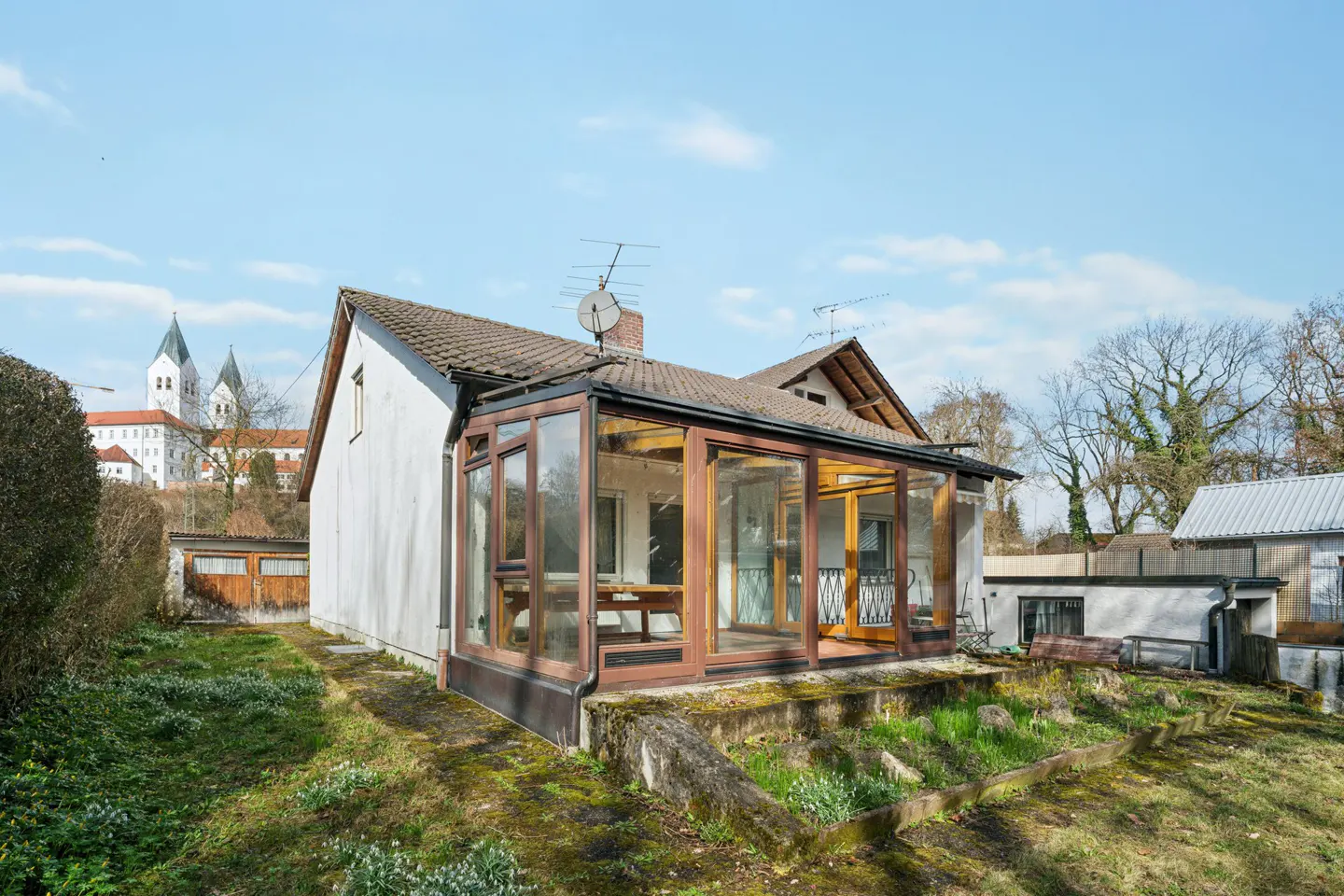 Exterior view of a white house with a glass sunroom addition and a grassy yard. A church is visible in the background.