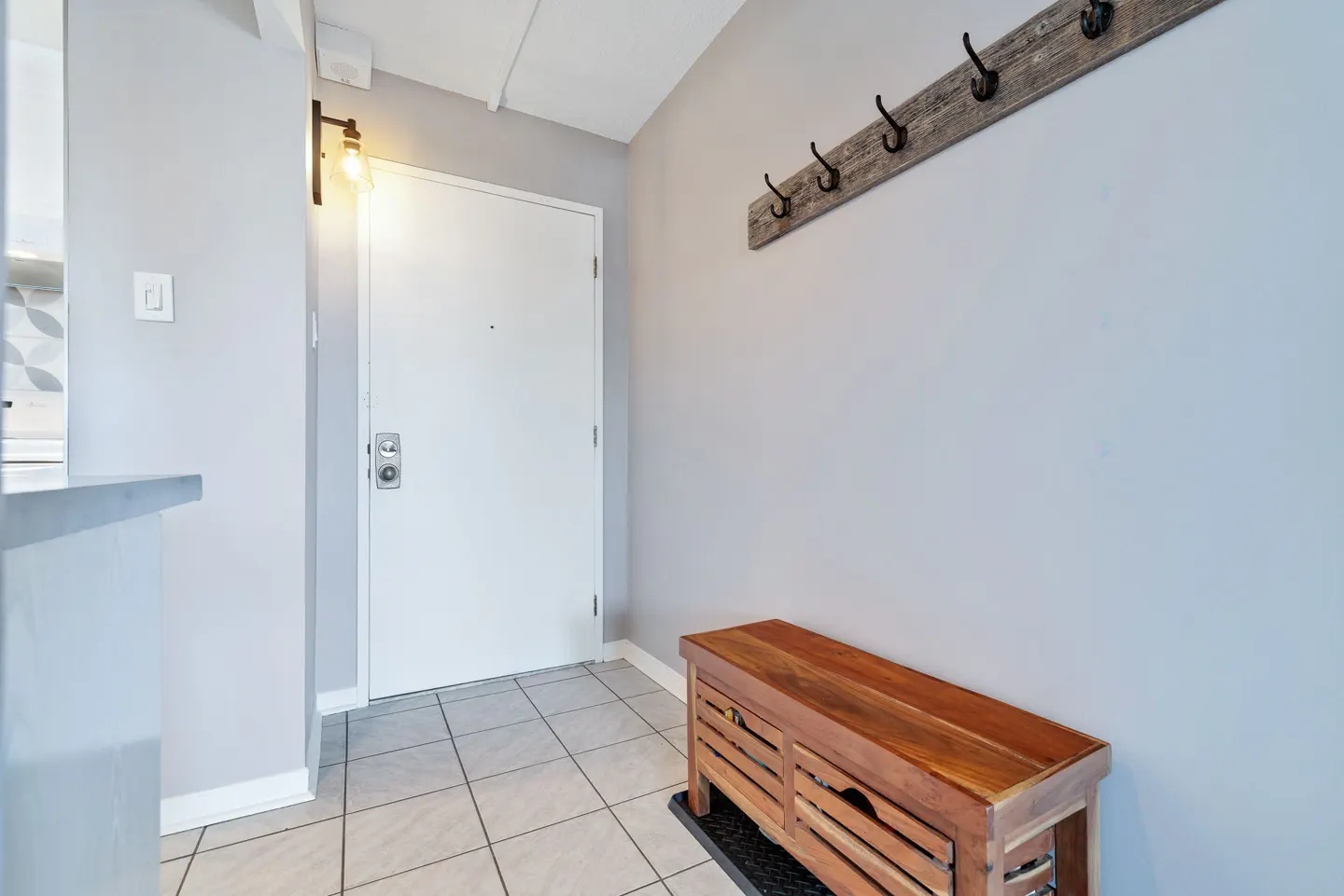 Entryway with a white door, light gray walls, and tiled floor. A wooden bench sits under a coat rack with black hooks.