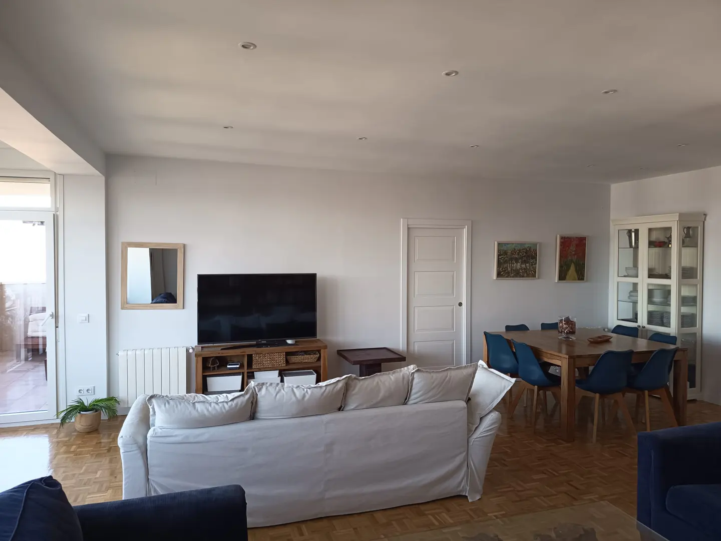 Spacious living room with a white sofa, TV on a wooden stand, and a dining area with blue chairs and a glass cabinet.