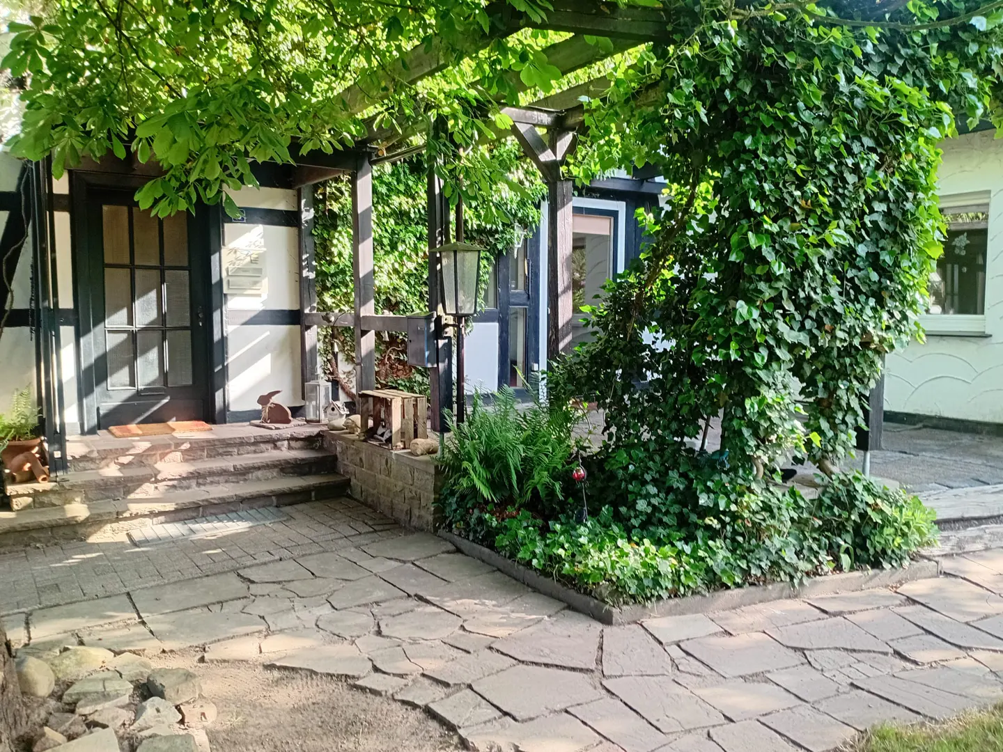 A stone patio leads to a half-timbered house entrance shaded by a lush green pergola covered in climbing ivy.