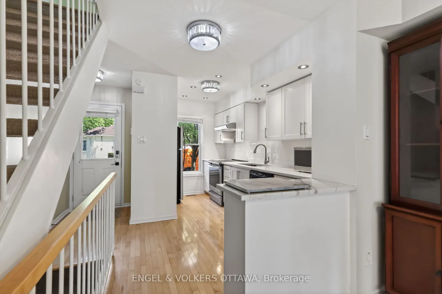 Bright, modern kitchen with white cabinets, stainless steel appliances, and hardwood floors. Staircase on the left, window at the end of the kitchen.