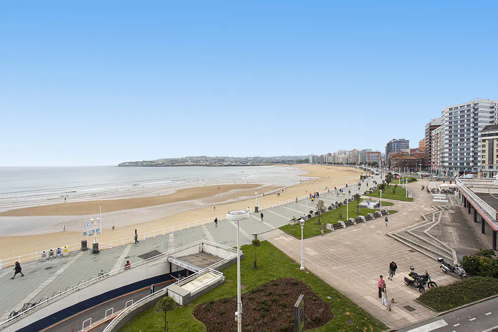 High-angle view of a sandy beach, a paved promenade with pedestrians, and city buildings under a clear blue sky.