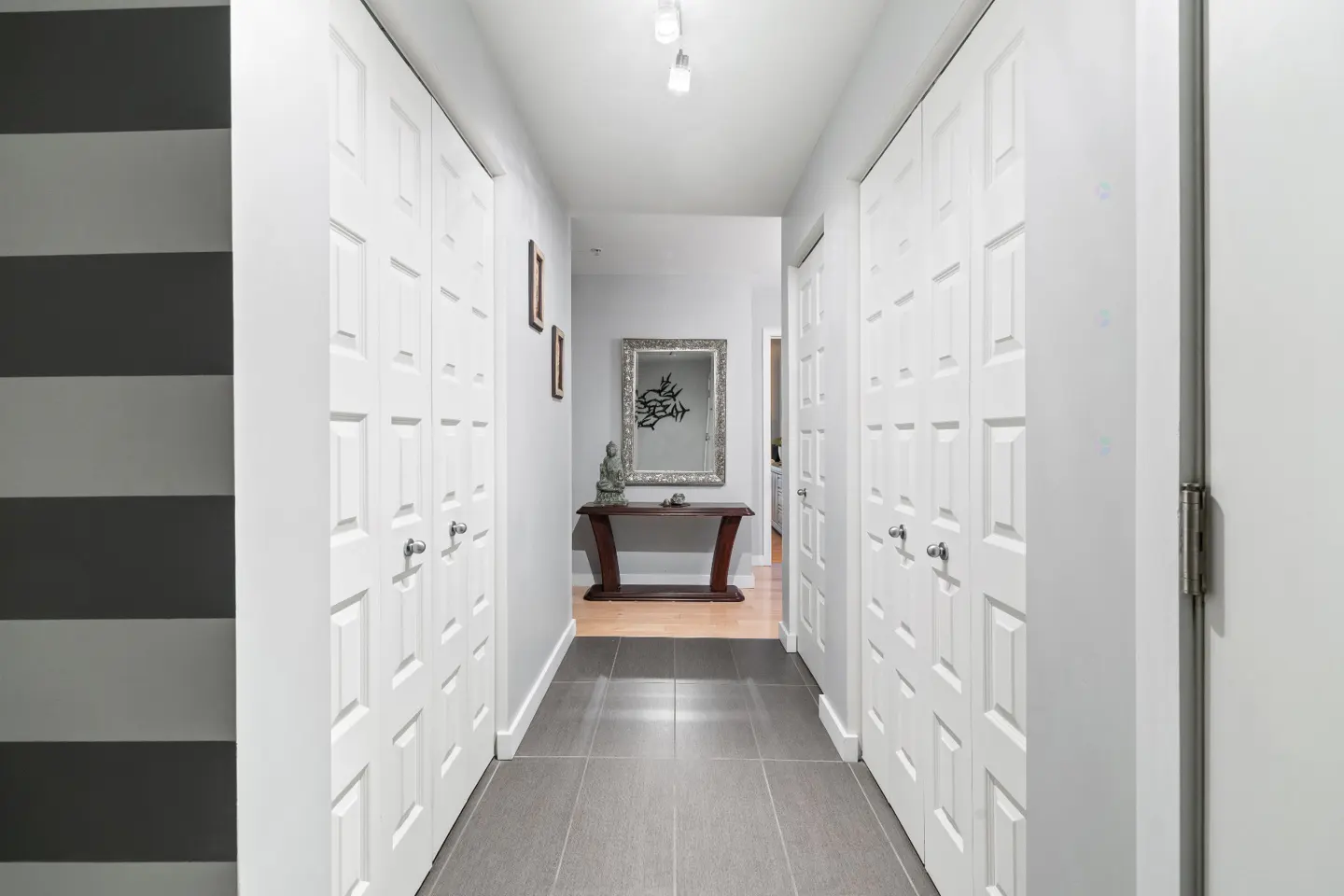 Hallway with gray tile floor, white walls, and white closet doors on both sides. A table with a mirror and statue is at the end.