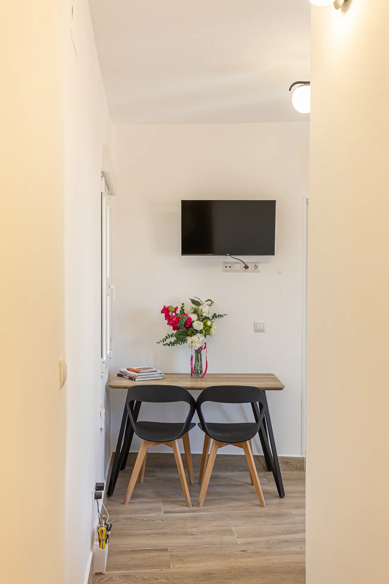 A dining area with a wooden table, two black chairs, a flower vase, and a wall-mounted television against a white wall.