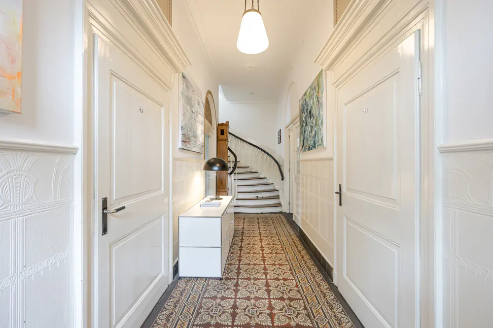 Hallway with white walls, doors, and patterned tile floor. A staircase is visible at the end of the hall. A white cabinet with a lamp sits to the left.