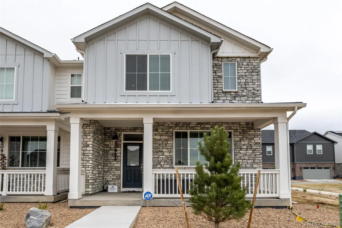 Two-story house with gray siding and stone accents. White porch columns and railing. A small pine tree is planted in the front yard.
