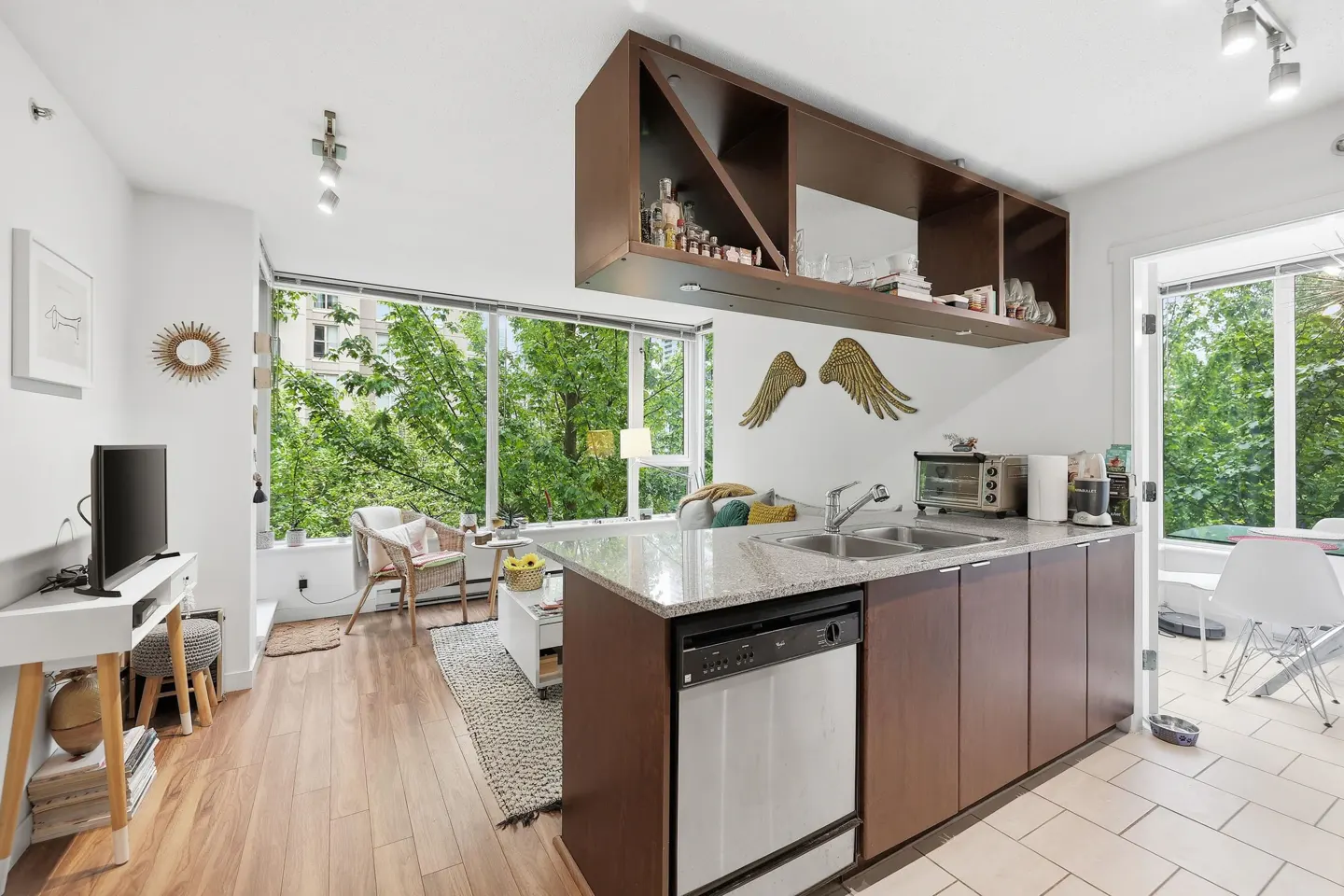 A bright, modern apartment interior with a kitchen island, wood floors, and a large window overlooking lush greenery.