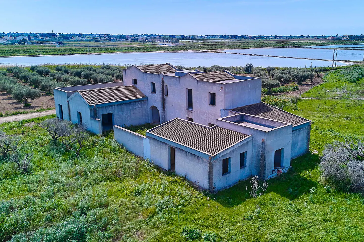 Aerial view of a large, pale blue, derelict building with a brown tiled roof, surrounded by green grass and trees, with a blue sky.