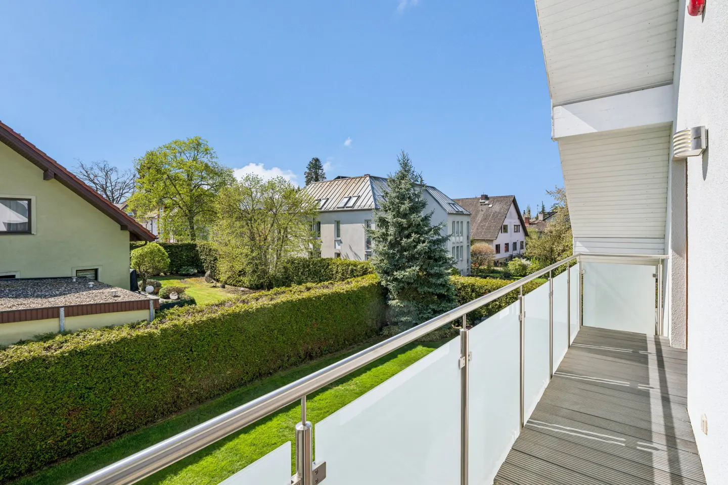View from a balcony with a metal and frosted glass railing, overlooking a green lawn, hedge, and houses under a blue sky.