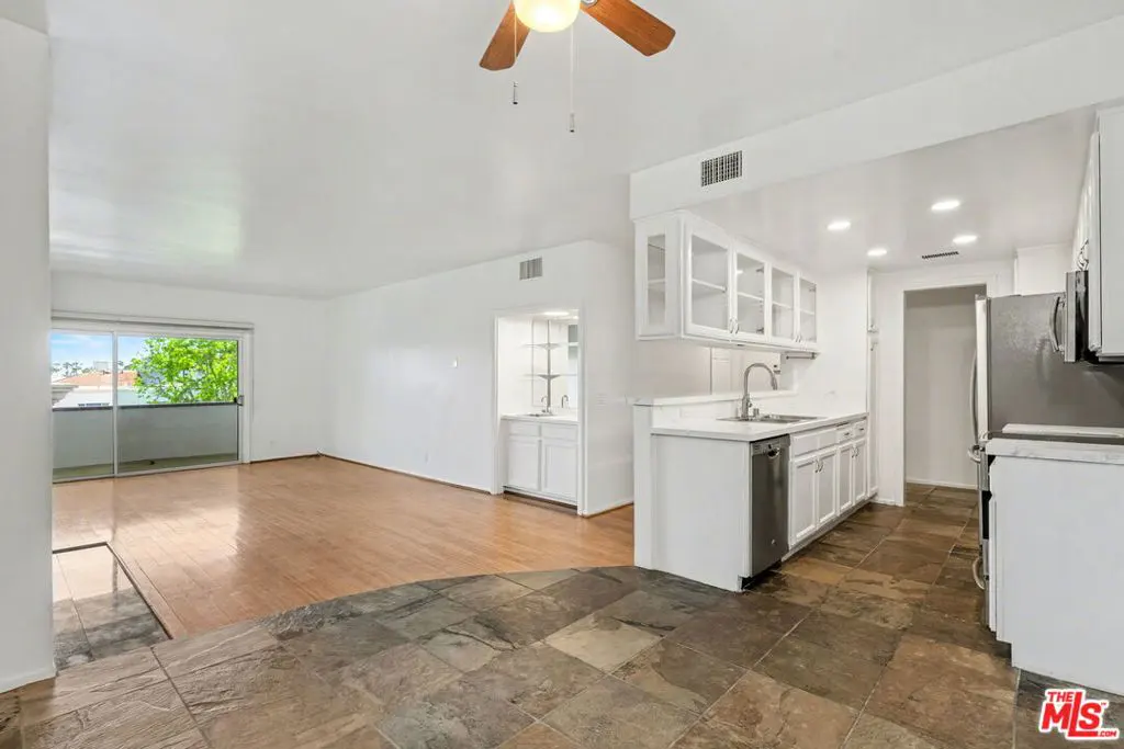 A bright, open-concept condo interior with white walls, wood floors, and a modern kitchen with white cabinets and stainless steel appliances. A balcony is visible.