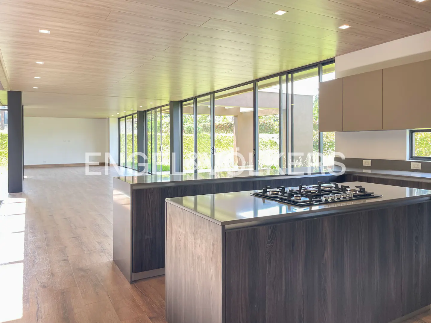 Open-concept kitchen with wood floors, a dark wood island with a gas cooktop, and floor-to-ceiling windows overlooking greenery.