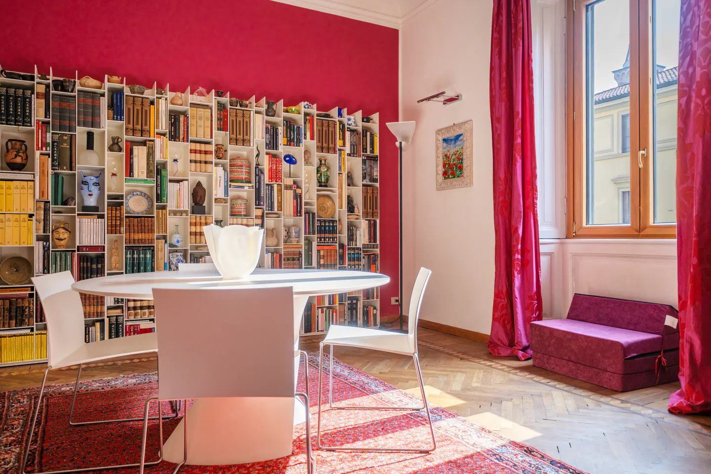 Bright dining room with a red accent wall, a large bookshelf, and a round table with white chairs. Red curtains frame a window with sunlight streaming in.